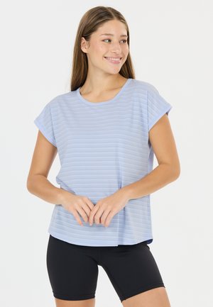 Ung kvinde smilende, iført en lyseblå stribet kortærmet skjorte og sorte cykelshorts, stående med hænderne sammen foran.