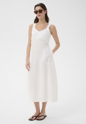 Femeie purtând o rochie midi albă fără mâneci, ochelari de soare închiși la culoare și șlapi negri, stând cu o mână în buzunar pe fundal alb.