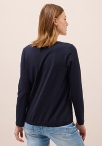 Marineblaues Langarmshirt mit rundem Ausschnitt, aus weichem Baumwollmaterial, lockere Passform, der hintere Saum ist etwas länger als vorne, getragen mit hellblauen Jeans.