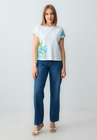Weißes T-Shirt mit kurzen Ärmeln und blauer Blumenmusterung auf der linken Seite, kombiniert mit hochgeschnittenen blauen Jeans und weißen offenen Sandalen.