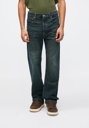 Homme portant un jean droit bleu foncé, une chemise verte et des baskets marron, debout sur un fond uni.