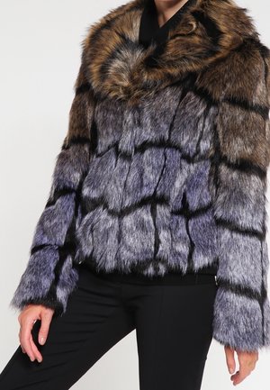 Frau trägt eine mehrfarbige Pelzjacke mit braunen, grauen und schwarzen Streifen, kombiniert mit schwarzen Hosen vor einem schlichten Hintergrund.