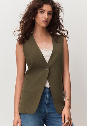 Ung kvinde med krøllet brunt hår iført olivengrøn ærmeløs blazer over hvid top med blonder og blå jeans, stående mod en ensfarvet baggrund.