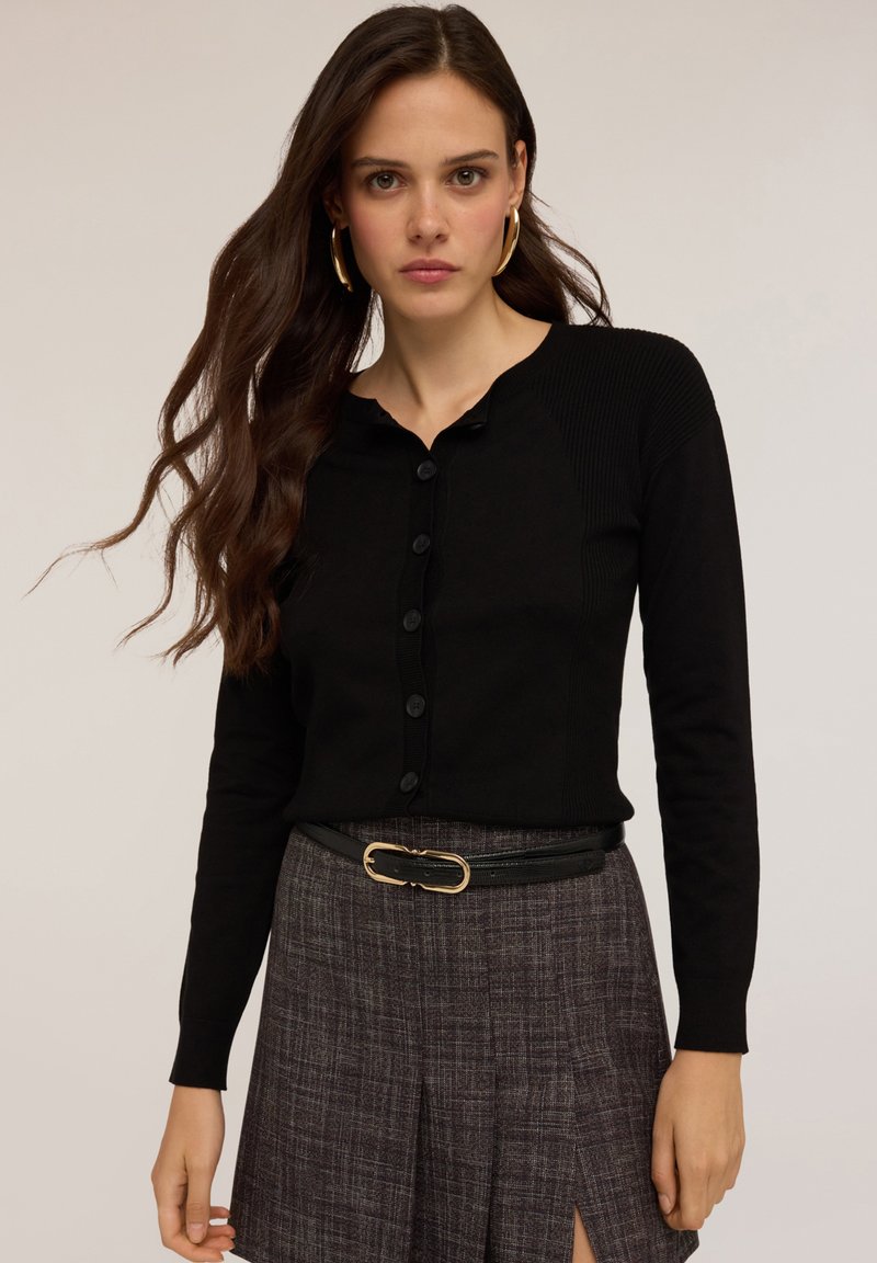 Svart långärmad kavaj med knäppning fram, ribbad textur och rundad halsringning, kombinerad med en rutmönstrad kjol och ett svart bälte.