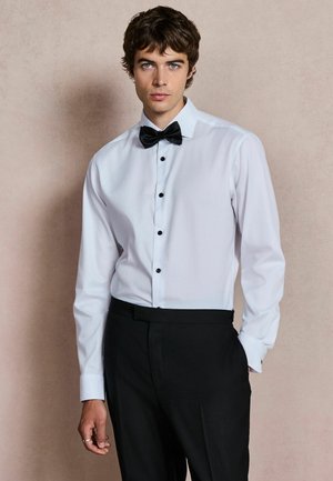 Joven con cabello oscuro y rizado, vestido con una camisa blanca, pajarita negra y pantalones negros sobre un fondo liso beige.