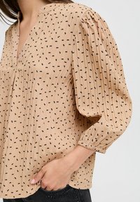 Blouse beige à manches longues avec un décolleté en V, présentant un tissu texturé avec de petits motifs de points noirs et des manches froncées. Plis subtils à l'avant.