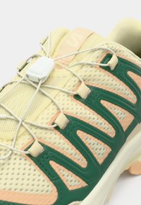 Αθλητικό παπούτσι σε ανοιχτό κίτρινο χρώμα με πράσινες και ροδακινί λεπτομέρειες, επάνω μέρος από δίχτυ με έντονες ραβδώσεις, ελαστικό σύστημα κορδονιών και υφή στη σόλα.