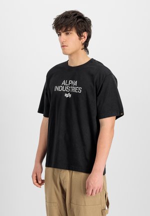 Young man with dark hair wears black "Alpha Industries" T-shirt and beige cargo pants, standing against plain light background.