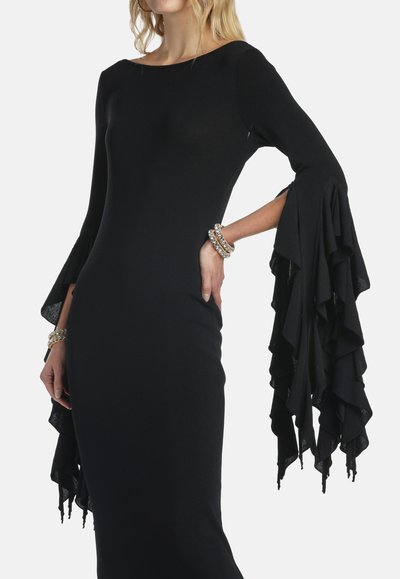 Mujer con un vestido negro ajustado que presenta mangas largas con volantes en capas y lleva pulseras doradas en ambas muñecas.