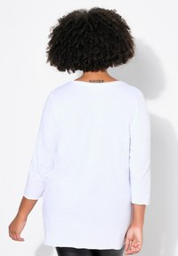 Top bianco a maniche lunghe con scollo rotondo, vestibilità rilassata e tessuto leggermente strutturato. La vista dal retro mostra un design semplice e un taglio più lungo.