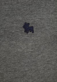 Piccolo cane ricamato di un blu navy su tessuto grigio testurizzato, centrato su una superficie tessile.