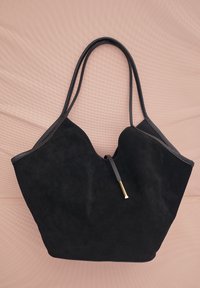 Melnas zamšādas somiņas ar divām rokturiem un izteiksmīgu izliekumu. Uzsvars uz zelta toņu aksesuāriem. Gluda, teksturēta fona.