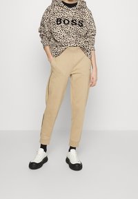 Leopardenmuster-Hoodie mit "BOSS" Schriftzug, beigem Sweatpants mit seitlichen Streifen und schwarz-weißen klobigen Turnschuhen.