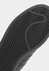 Suola di scarpa in gomma nera con un motivo di battistrada ondulato, caratterizzata da creste e scanalature per una migliore aderenza. Bordo liscio e superficie leggermente testurizzata.