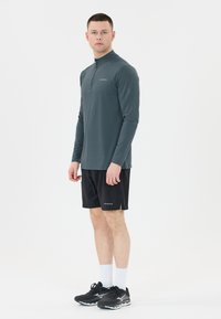 Dunkelgraues Langarmshirt mit halbem Reißverschluss, kombiniert mit schwarzen Sportshorts. Schwarze Sneaker und weiße Socken vervollständigen das Outfit.
