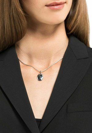 Collana d'argento con un pendente geometrico in metallo nero lucido, indossata su una camicia nera con colletto. Primo piano del pendente e dei dettagli della catena.