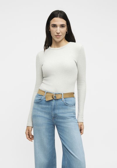 Νεαρή γυναίκα με μακριά σκουρόχρωμα μαλλιά που φοράει ένα ανοιχτό γκρι εφαρμοστό μακρυμάνικο μπλουζάκι, ανοιχτά μπλε ψηλόμεσα τζιν και μια καφέ ζώνη.