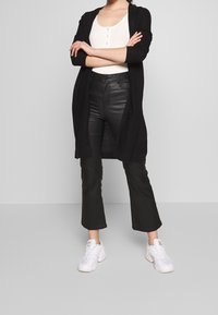 Cardigan noir long porté sur un haut blanc côtelé, associé à un pantalon flare en cuir noir taille haute et des baskets blanches.