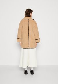 Beżowy, puszysty płaszcz z czarną lamówką, o luźnym kroju, szerokich rękawach i prostym fasonie, zestawiony z białymi spodniami o szerokich nogawkach oraz czarnymi butami.