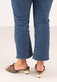 Sandalias de slide tejidas en negro con un chunky tacón bloque dorado, combinadas con unos vaqueros cortos azules con dobladillos deshilachados.