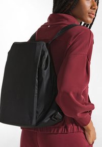 Zaino in tessuto nero di forma rettangolare, con una tasca frontale e spallacci regolabili, indossato sopra un outfit color bordeaux.