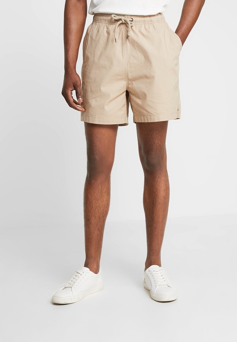 Homme portant un short beige à cordon de serrage et des baskets blanches, debout avec une main dans la poche sur un fond neutre.