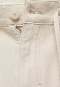 Gros plan sur la ceinture d'un pantalon beige montrant un bouton, une fermeture éclair partiellement ouverte et une boutonnière cousue sur le tissu.