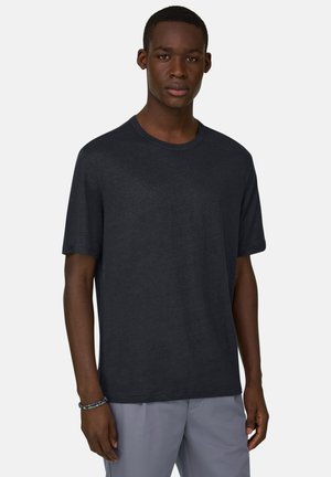 Junger Mann, der ein schlichtes dunkelgraues kurzärmeliges T-Shirt und hellgraue Hosen trägt, vor einem neutralen Hintergrund stehend.