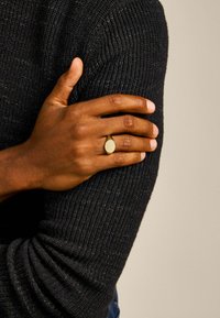 Gouden metalen ring met een ovale vorm, gedragen op een hand tegen een donkere, gestructureerde trui-achtergrond. Huidskleur is diep bruin.