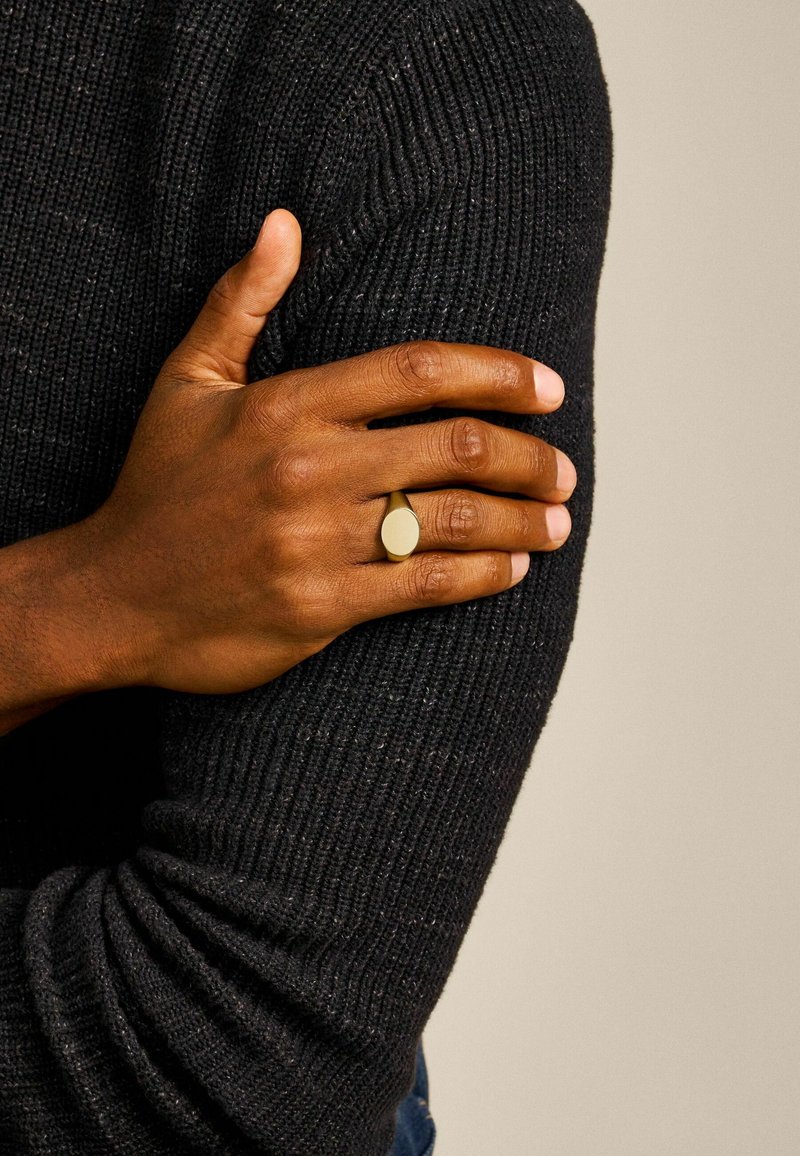 Gouden metalen ring met een ovale vorm, gedragen op een hand tegen een donkere, gestructureerde trui-achtergrond. Huidskleur is diep bruin.