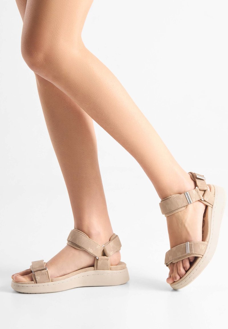Beige platåsandaler med justerbare remme, lavet af blødt materiale. Har flade såler, en tekstureret overflade og metalliske accenter.