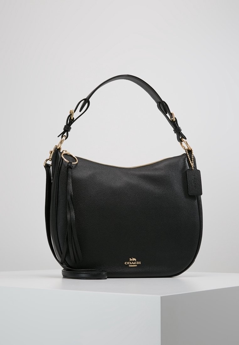 Sac à main Coach en cuir noir avec ferrures dorées, poignée supérieure, tirette à pompon et logo sur le devant, posé sur une surface blanche.
