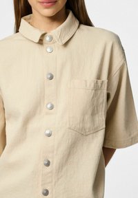 Bež majica s kratkimi rokavi iz bombažnega materiala, z gumbnim zapiranjem spredaj, srebrnimi gumbi, ovratnikom in majhnim žepkom na prsih.