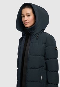Chaqueta acolchada de color azul oscuro con una gran capucha, cierre con cremallera, dos bolsillos laterales y un detalle de logo en la manga. Tejido texturizado con un acabado suave.