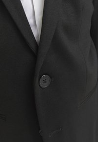 Detailný záber na čierne sako zapnuté cez bielu košeľu, zvýrazňujúci textúru látky a jeden čierny gombík.