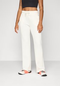 Een persoon staat in een zwart crop top en crèmekleurige sweatpants, gecombineerd met kleurrijke sportschoenen. De achtergrond is neutraal.