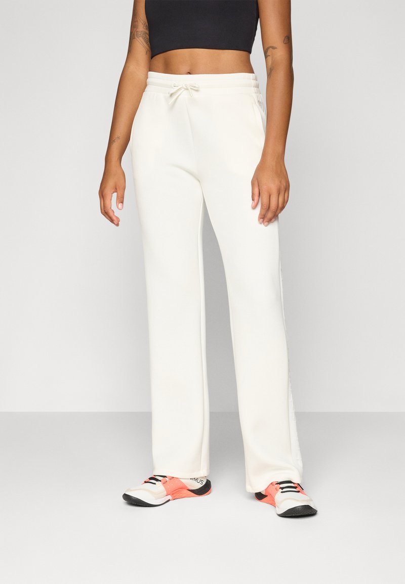 Een persoon staat in een zwart crop top en crèmekleurige sweatpants, gecombineerd met kleurrijke sportschoenen. De achtergrond is neutraal.