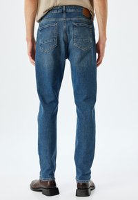 Jeans de mezclilla azul con corte recto, cinco bolsillos y un parche de cuero marrón en la cinturilla. Lavado medio con una textura sutil.