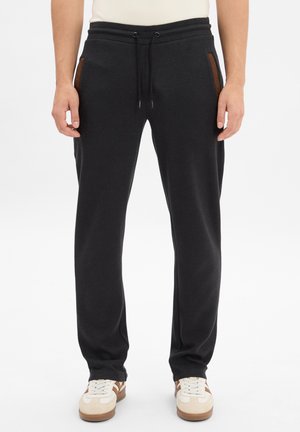 Schwarze Sweatpants mit einem Kordelzugbund, Seitentaschen mit braunen Akzenten und einem entspannten Schnitt. Getragen mit beigen und braunen Sneakers.