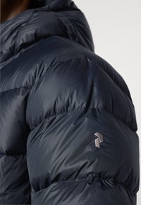 Blouson matelassé bleu marine avec une texture brillante et des sections piquées ; comporte un petit logo argenté sur la manche gauche.