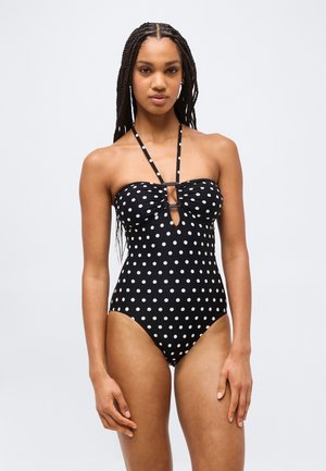 Femme aux longues tresses portant un maillot de bain noir à pois sans bretelles avec des lanières nouées au cou et une découpe à l'avant, se tenant devant un fond uni.