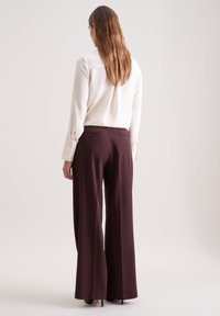 Crème blouse met knoopmanchetten en een rugstuk, gecombineerd met burgundy broek met wijde pijpen en dubbele achterzakken. Glad textuur.