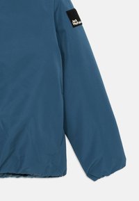 Modra nepremočljiva jakna z mat teksturo, z velcro nalepko na rokavu z napisom "Jack Wolfskin". Ohlapen kroj z elastičnimi manšetami.