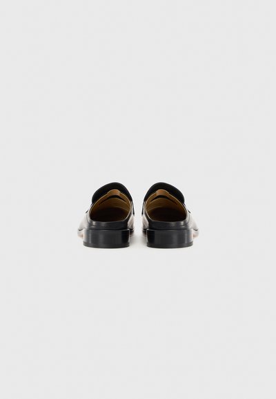 Ett par svarta läderloafers sedd bakifrån, som visar låga klackar och beige innersula mot en enkel vit bakgrund.