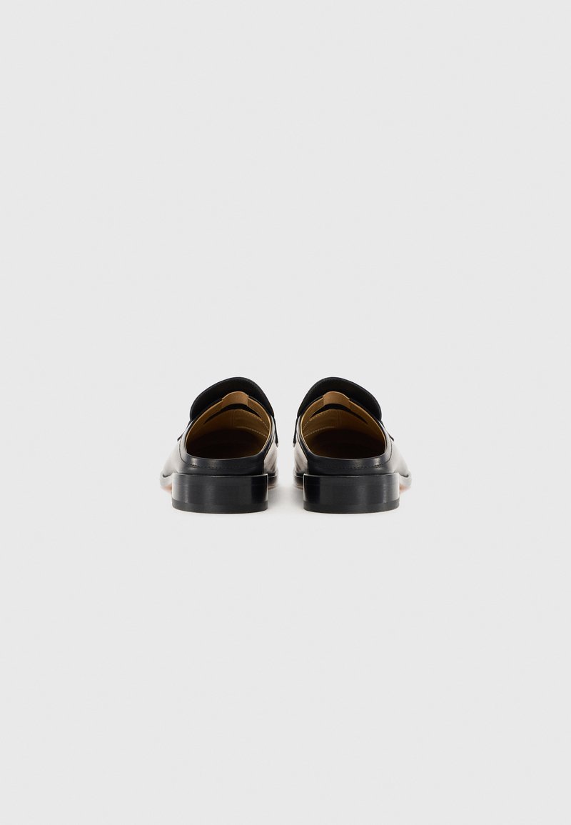 Ein Paar schwarze Lederloafer, von hinten gesehen, mit niedrigen Absätzen und einem beige Innenfutter vor einem schlichten weißen Hintergrund.