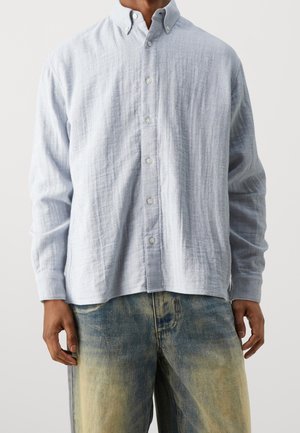 Homme portant une chemise boutonnée bleu clair texturée et un jean délavé bleu et beige, se tenant avec les bras détendus le long du corps.