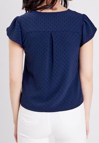 Tummaa sininen lyhythihainen paita, jossa on tekstuuria, takana laskos ja pyöreä pääntie. Yhdistetty valkoisiin housuihin.