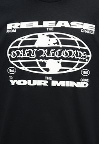 Μαύρο πουκάμισο με λευκό γραφικό κείμενο «Απελευθέρωσε το μυαλό σου από τη κούνια μέχρι τον τάφο» και οβάλ σχέδιο γης με το κείμενο «Obey Records» σε στιλιζαρισμένη γραμματοσειρά.