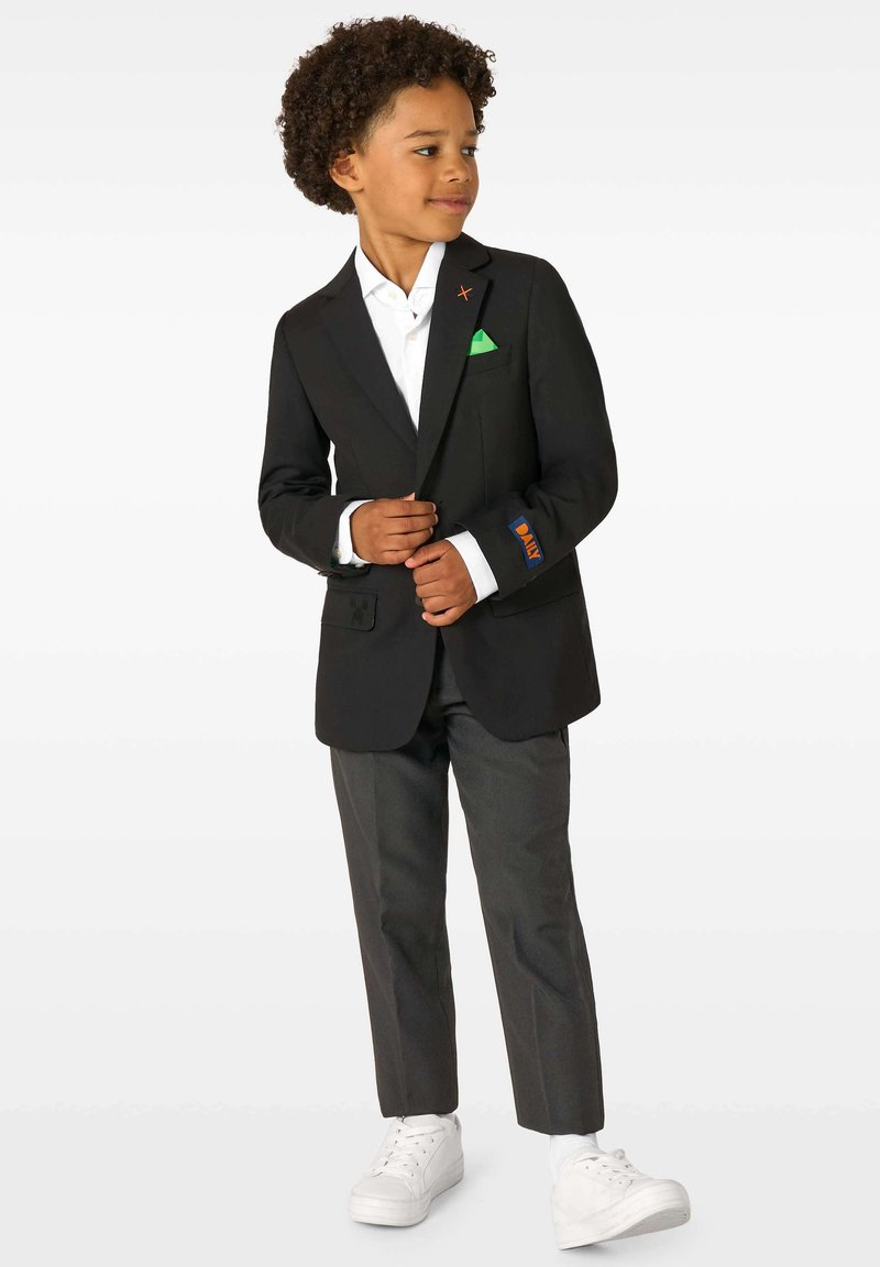 Kurruliste juustega poiss, kes kannab musta bleiserit valge särke ja tumedate pükste kohal, paigaldab bleiseri nööpe, seistes valge tausta ees.