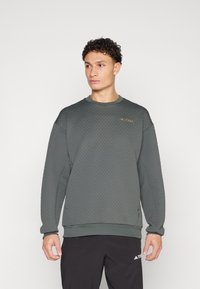 Sweatshirt texturé vert foncé avec un col rond et des poignets côtelés, présentant un petit logo doré sur le haut de la poitrine gauche.